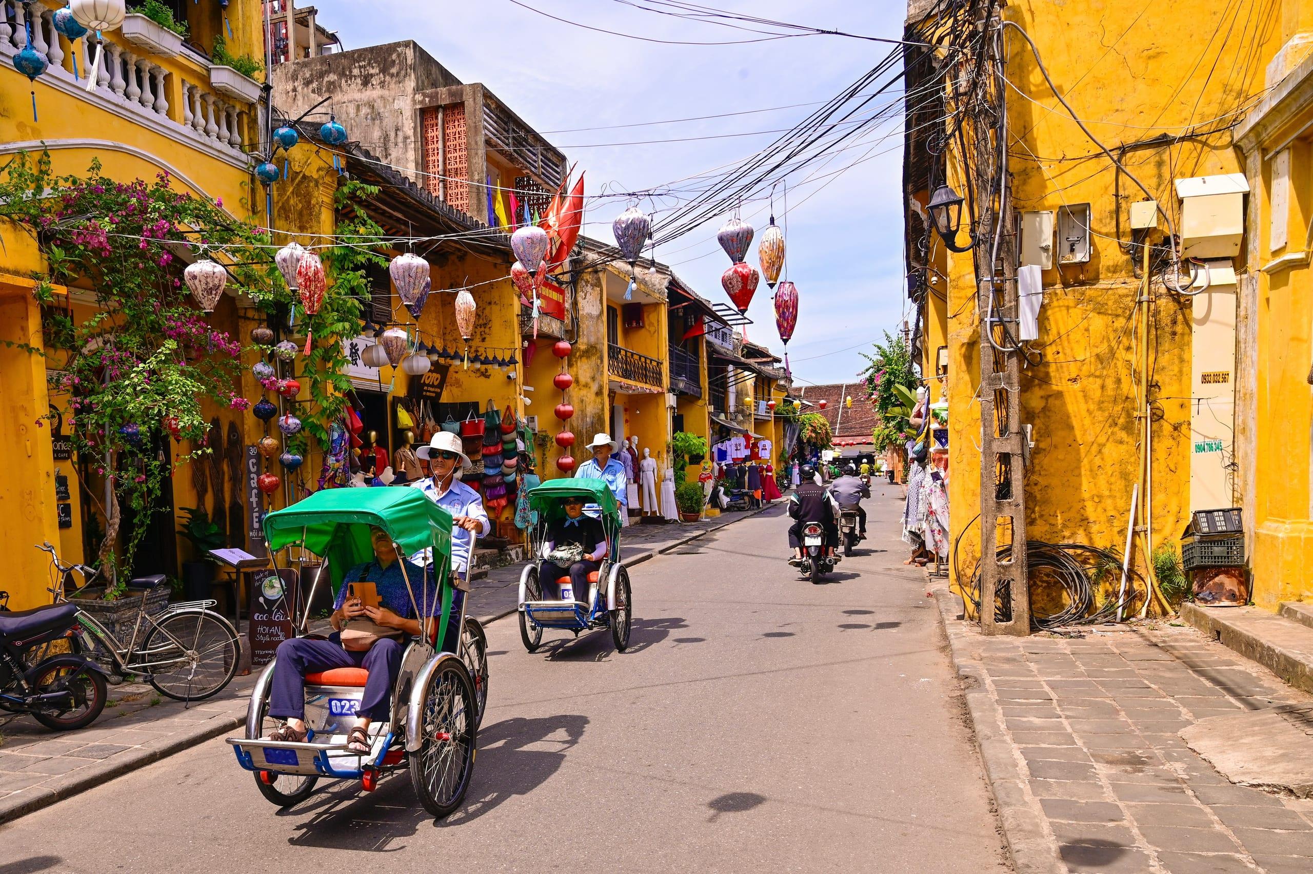Conducenti di risciò a pedali trasportano passeggeri lungo una strada gialla addobbata di lanterne a Hoi An