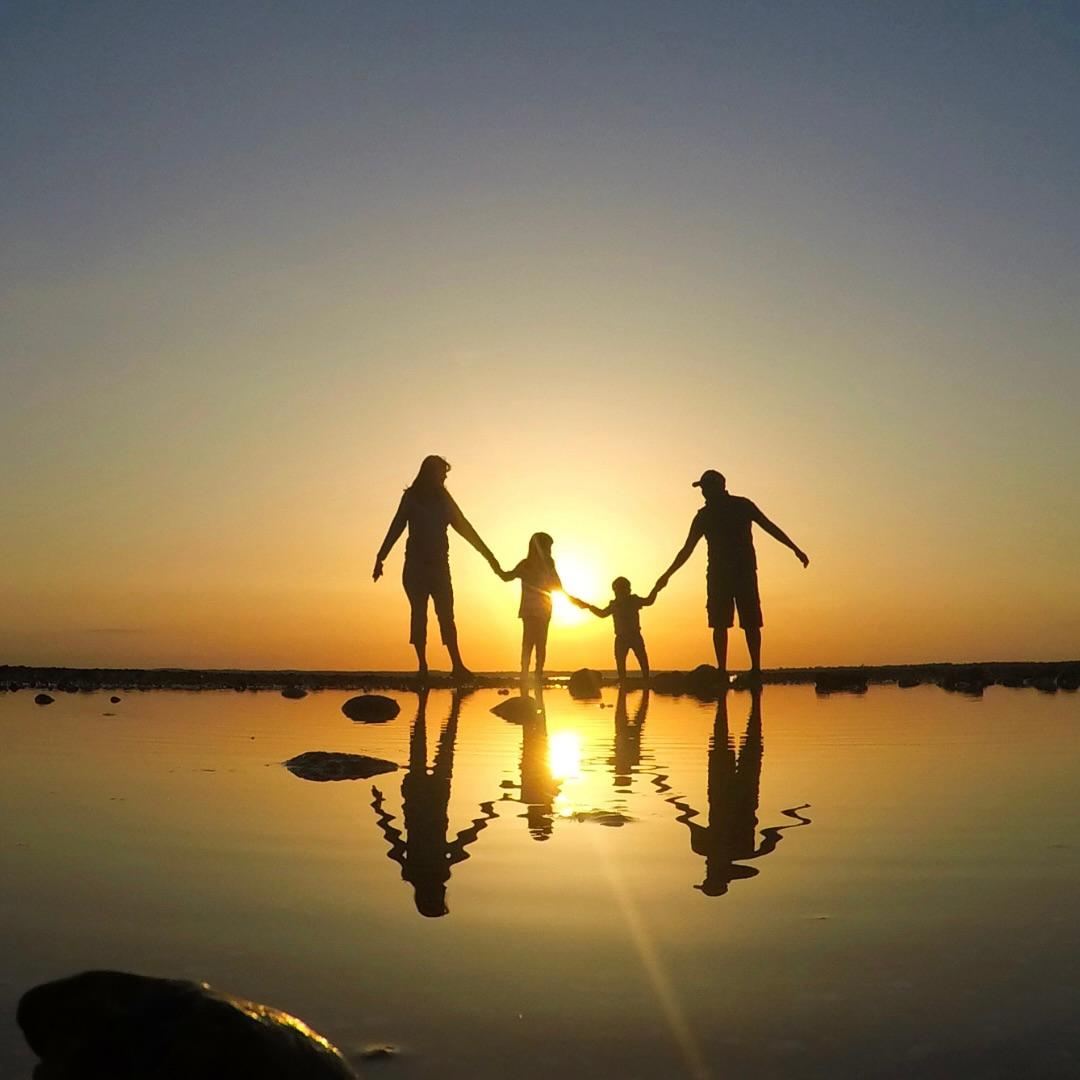 Una famiglia di quattro persone mano nella mano, in controluce al tramonto e riflessa nell’acqua bassa