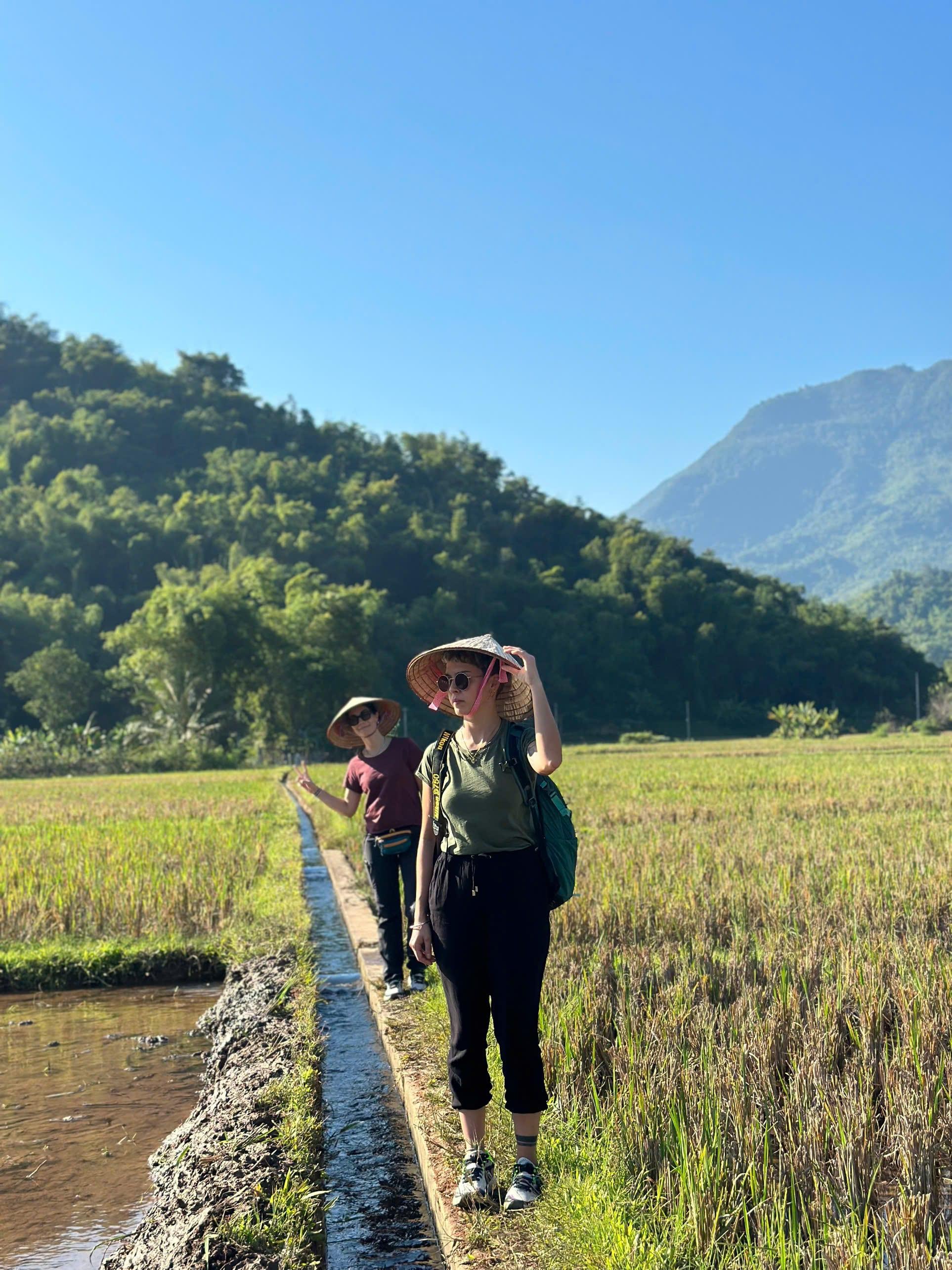 Due viaggiatori con il cappello conico percorrono uno stretto sentiero tra le risaie, sotto colline ricoperte di foreste