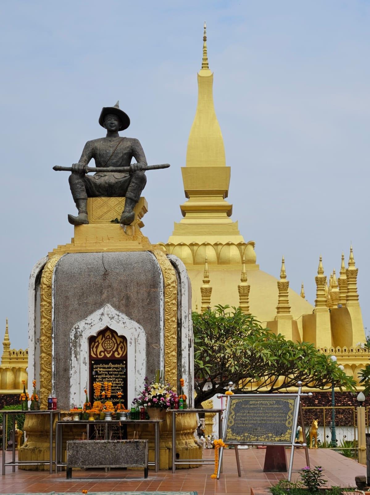 Uno stupa dorato che si innalza dietro la statua di un sovrano, con offerte alla base