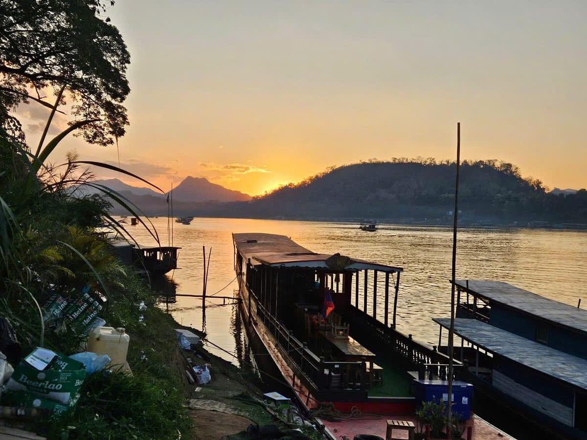 Barche di legno ormeggiate sul Mekong al tramonto, con le colline sulla sponda opposta