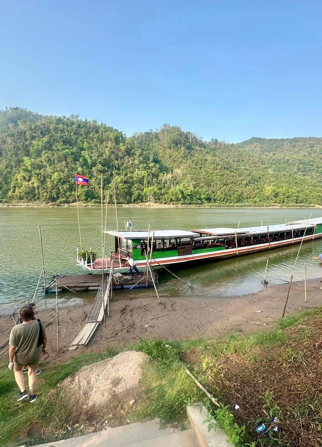 Una lunga barca fluviale ormeggiata alla riva, ai piedi di colline boscose