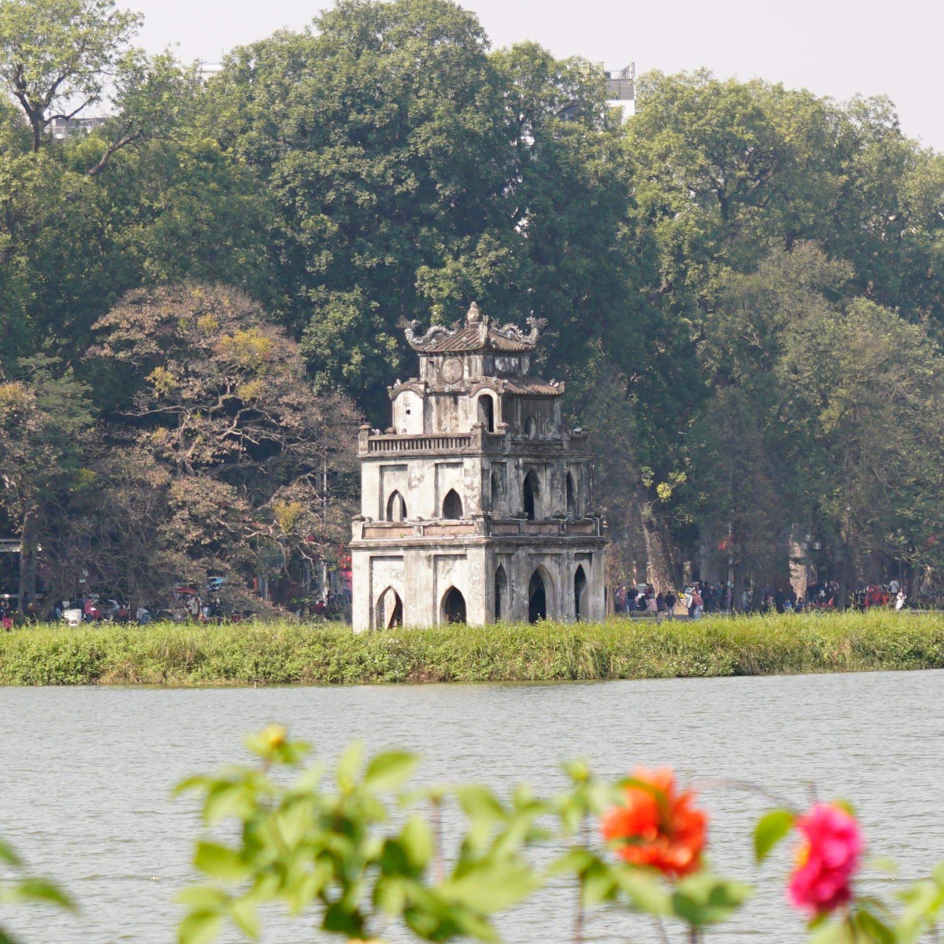 Una piccola torre di pietra su un isolotto di un lago cittadino, vista tra alberi e fiori