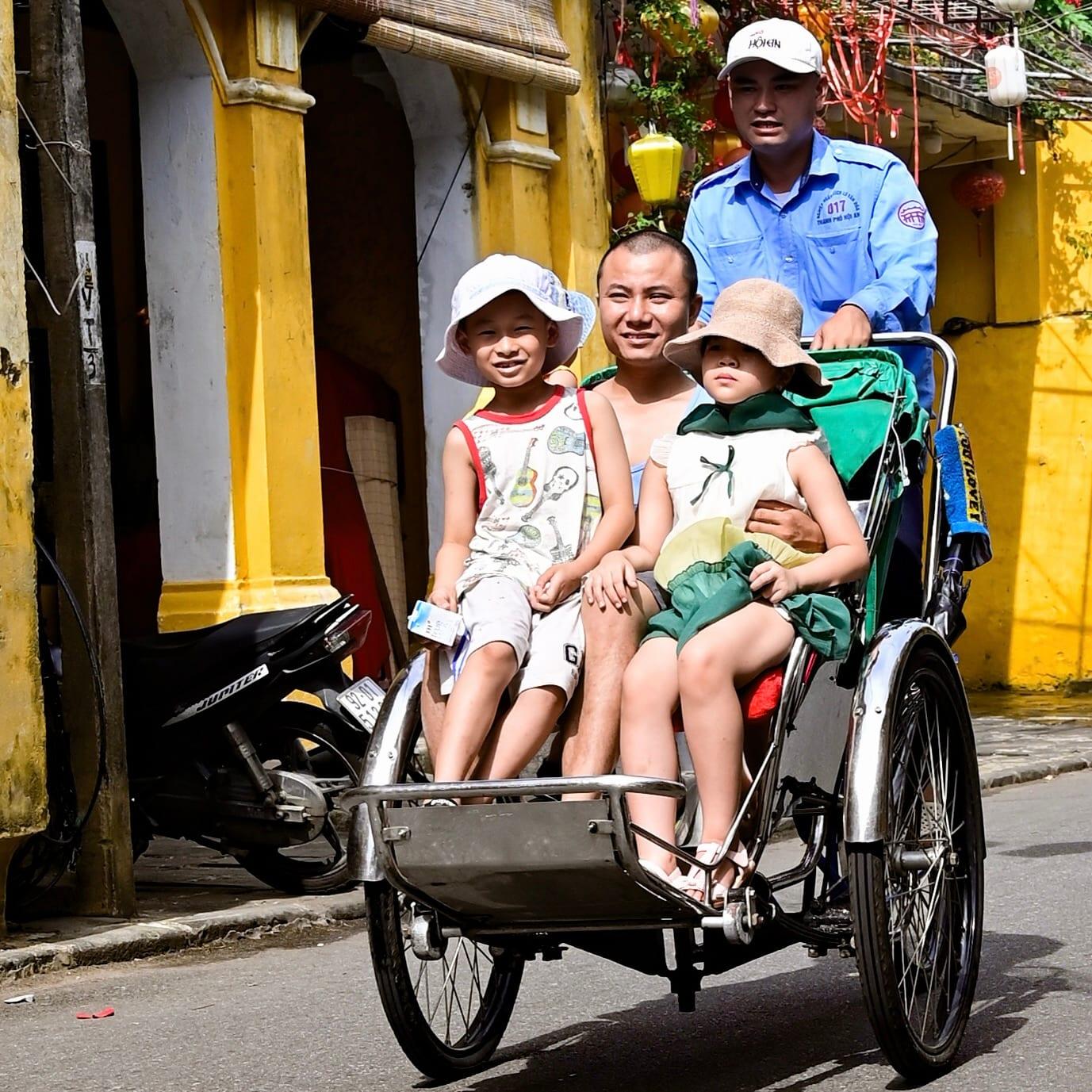 Due bambini con un adulto a bordo di un cyclo passano davanti a case-bottega gialle