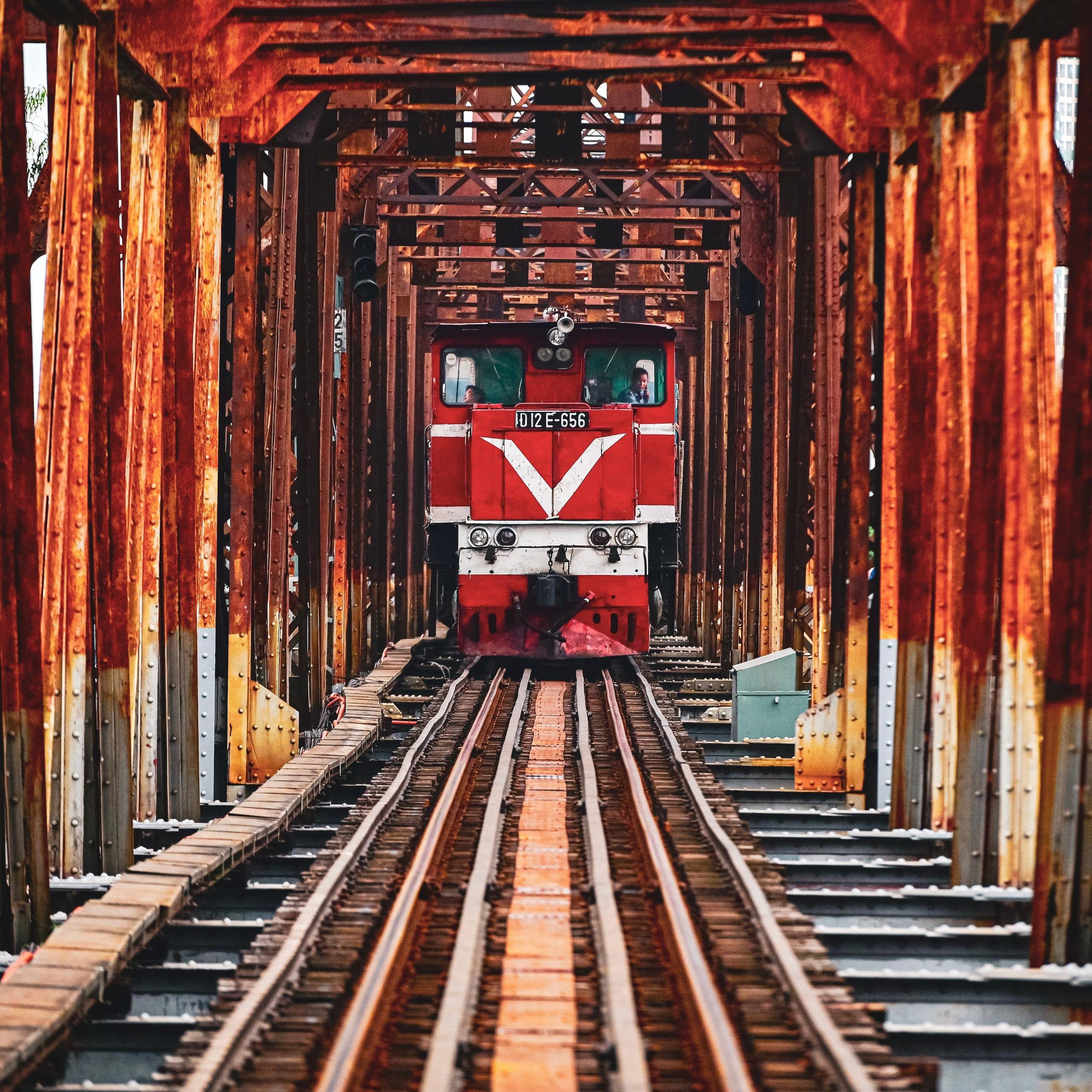 Una locomotiva rossa attraversa un vecchio ponte ferroviario in acciaio, vista frontalmente tra le travi